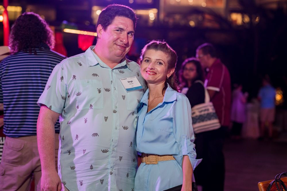 A man and woman smile together at an outdoor evening event, dressed in casual retro-style outfits.