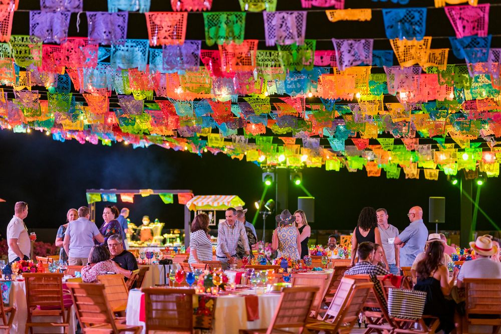 Colorful papel picado banners hang over festive outdoor Mexican fiesta dinner party with guests at decorated tables at night.