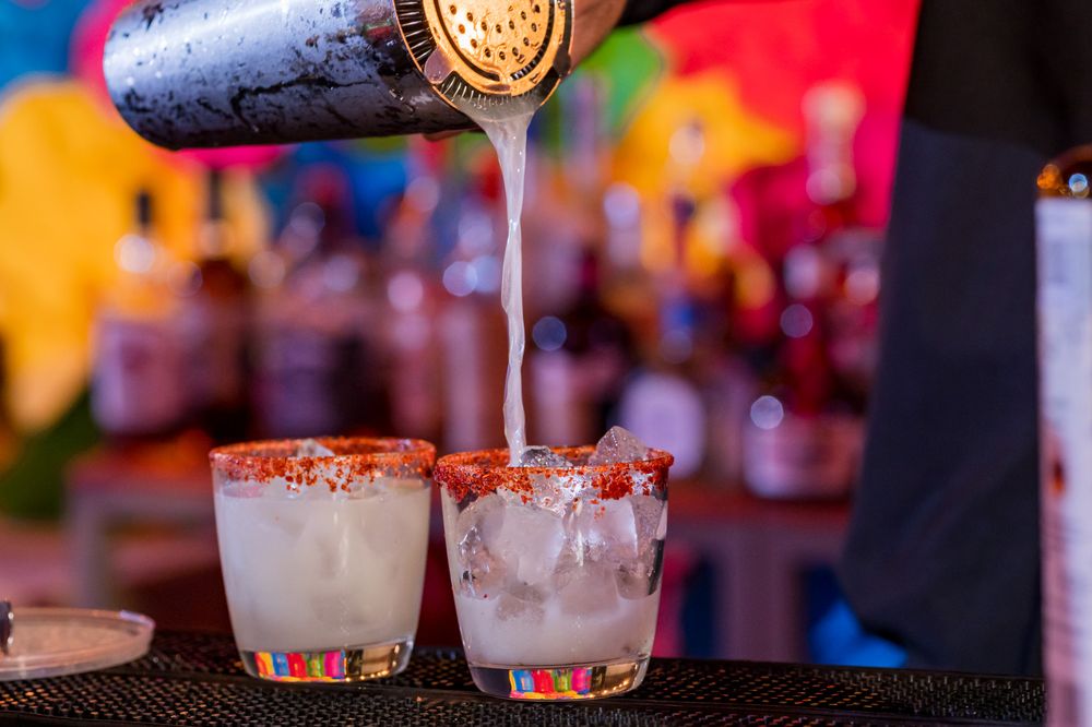 Bartender pouring margaritas into chili salt-rimmed glasses with ice at a colorful Mexican fiesta celebration.