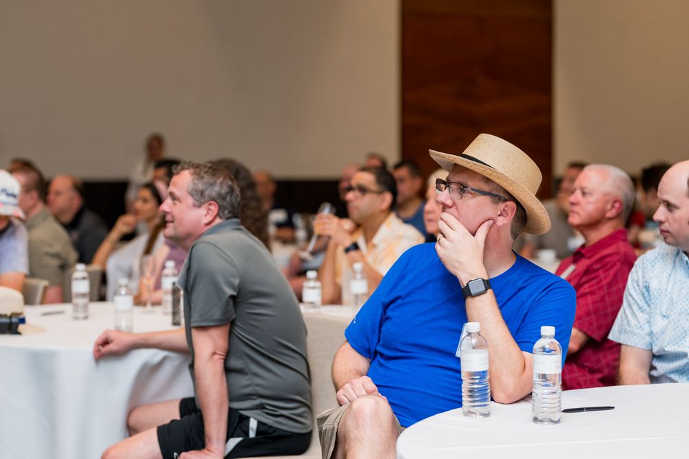 Men sitting at tables in a conference room, attentively watching a presentation, one wearing a straw hat and blue shirt.