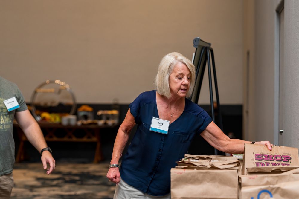 Woman in blue shirt moving brown paper bags at an indoor event or community gathering.