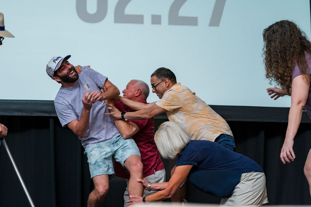 Three men grappling on a stage in front of a large screen displaying numbers at an outdoor event.