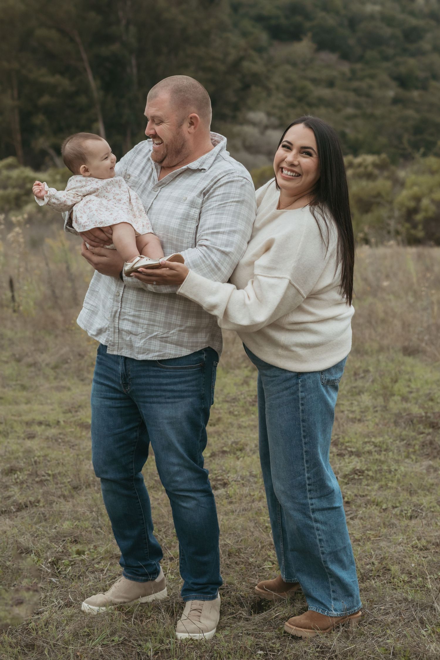 A happy family poses outdoors, with a man holding a baby and a woman smiling beside them in a grassy field.