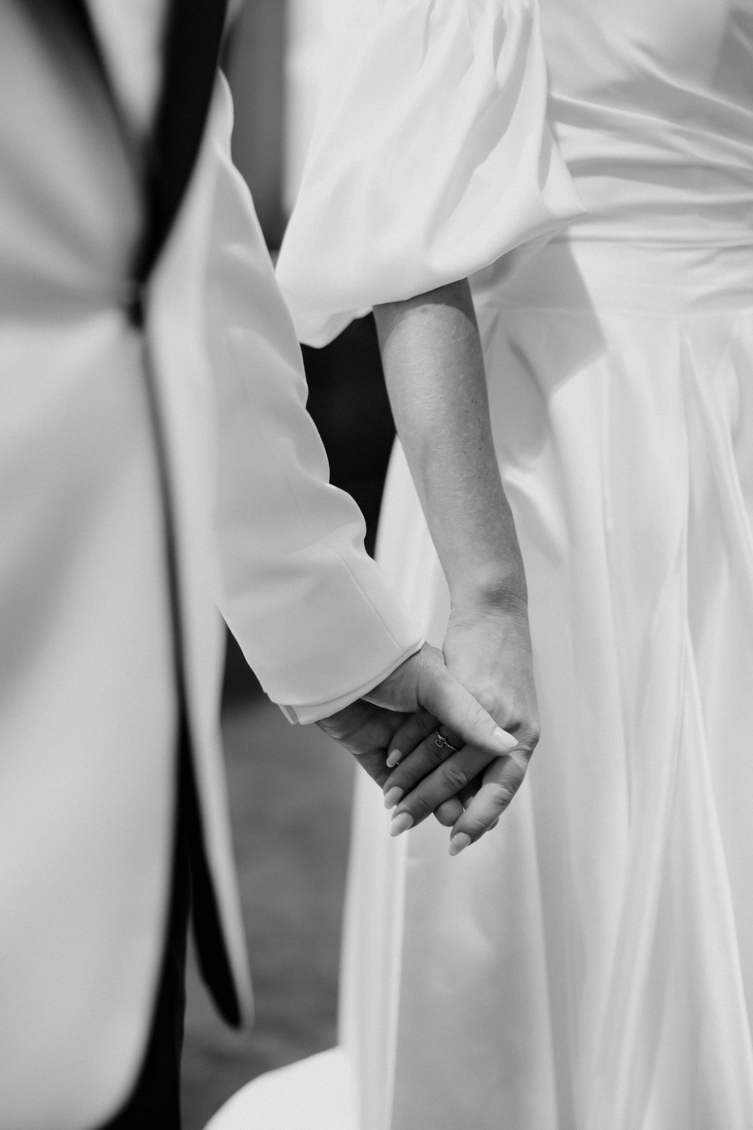 Black and white photo of a bride and groom holding hands, showcasing wedding rings and elegant white attire.