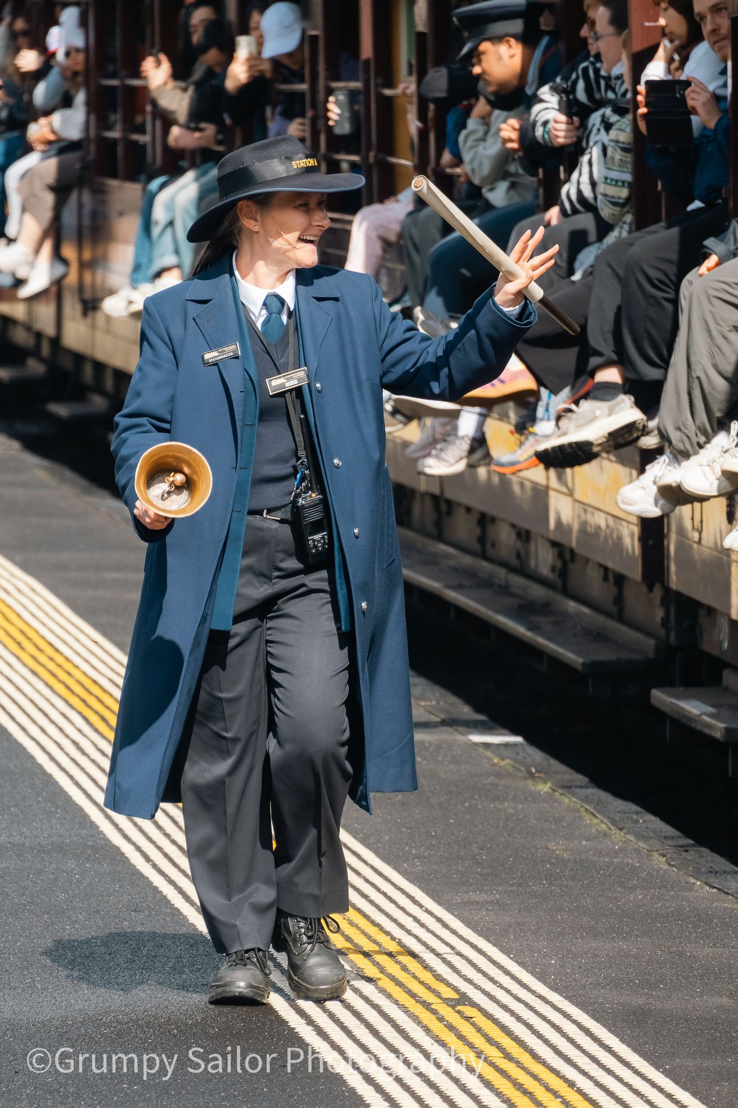 A train conductor in blue uniform holds a megaphone and waves on a busy station platform.