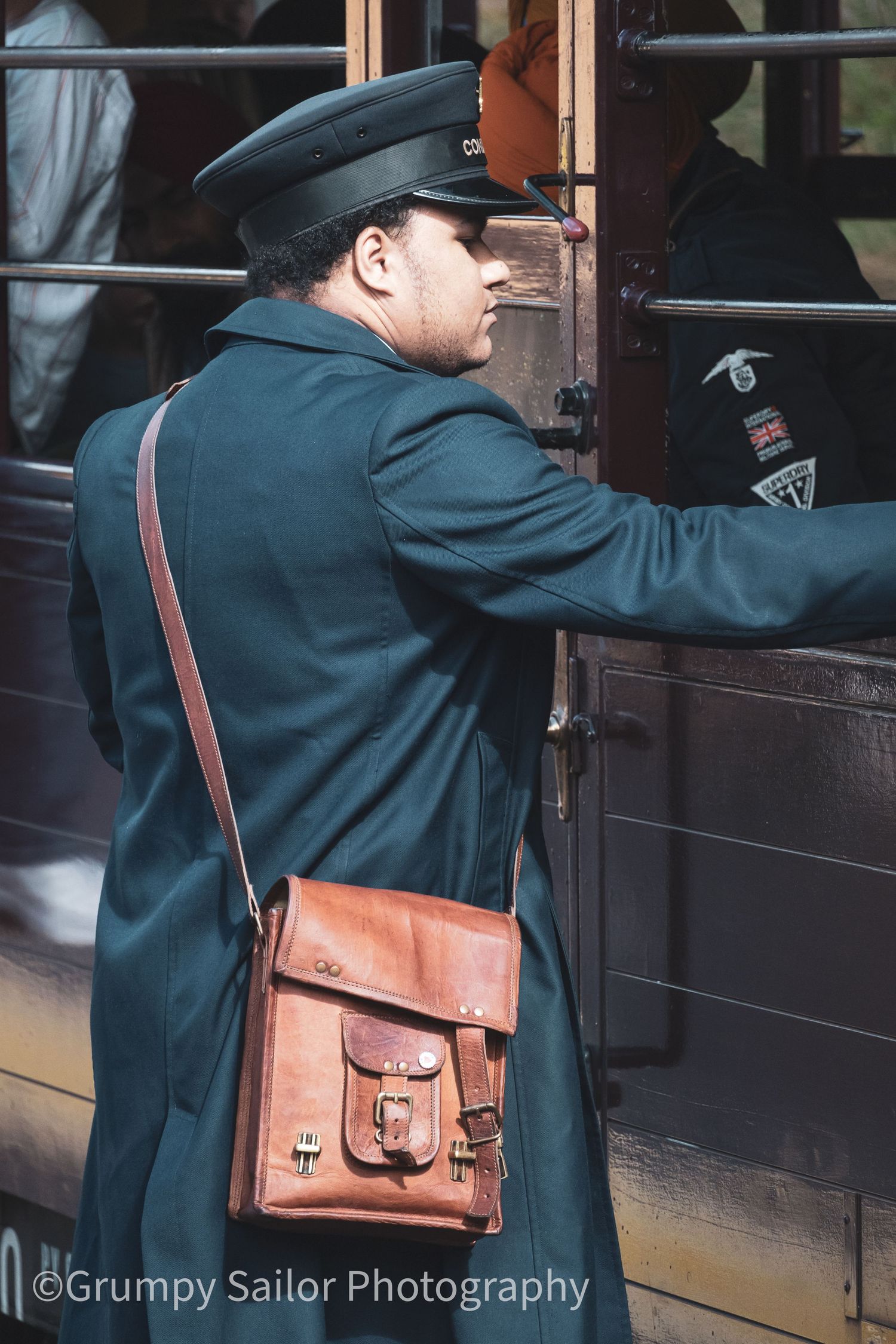 A uniformed conductor in a green coat and cap carries a brown leather satchel while boarding a vintage train.