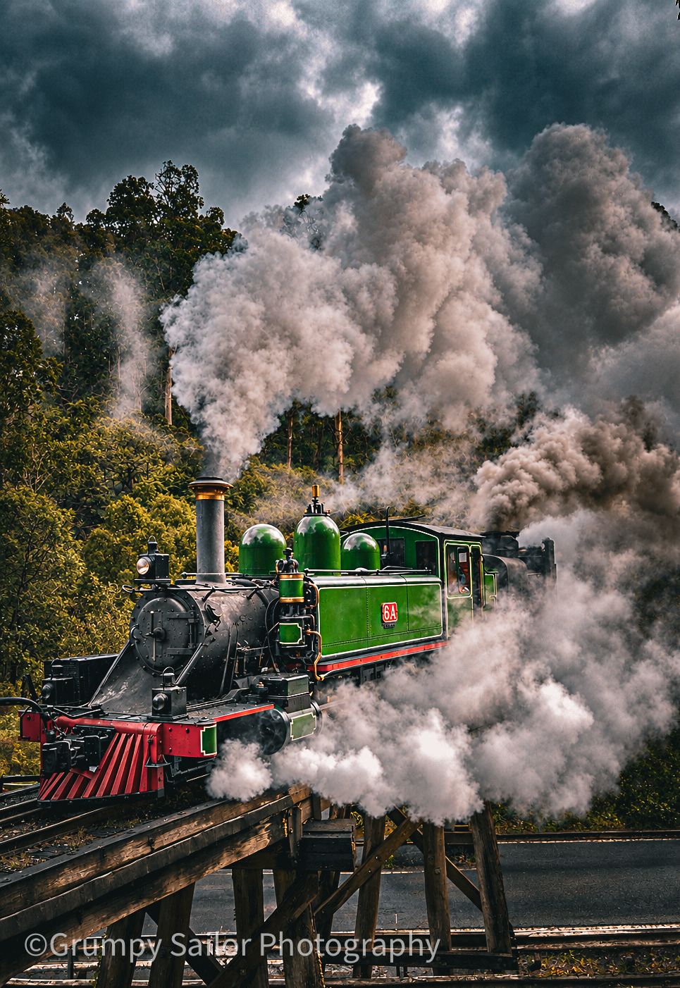 Vintage green steam locomotive billowing smoke on wooden trestle bridge under dramatic cloudy sky.