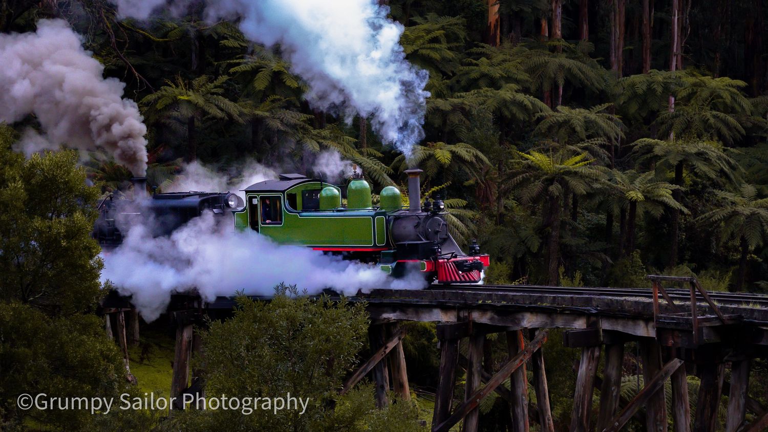 Vintage green steam locomotive crossing a wooden trestle bridge, billowing smoke, surrounded by lush forest trees.