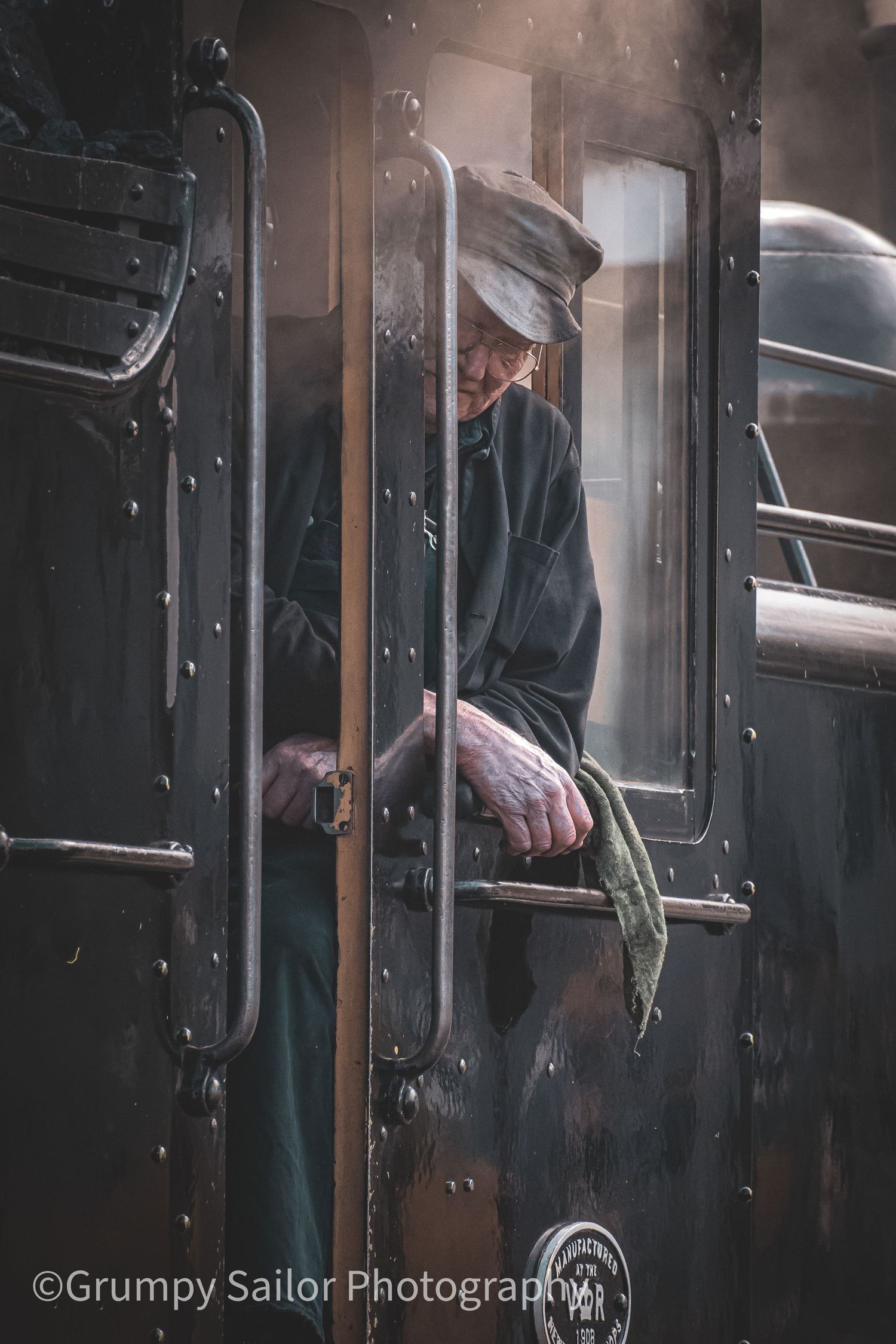 Elderly train conductor in vintage uniform leaning from dark steam locomotive door, holding rail with weathered hands.