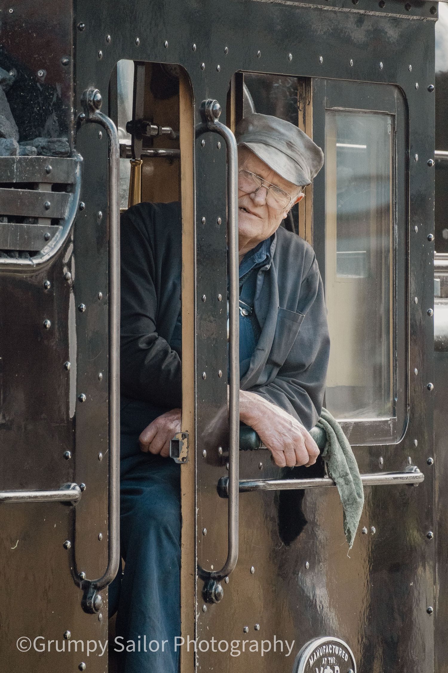 Elderly man in a flat cap and glasses leans out of a vintage steam train cab door, holding a cloth.