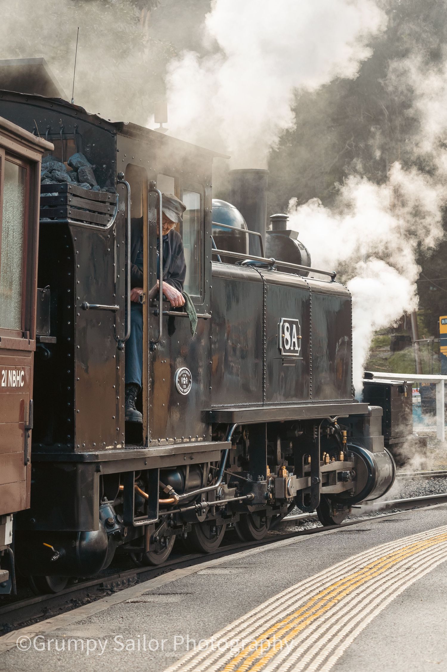 Vintage steam locomotive engine BA billowing white smoke at a railway station platform with engineer leaning out cab window.