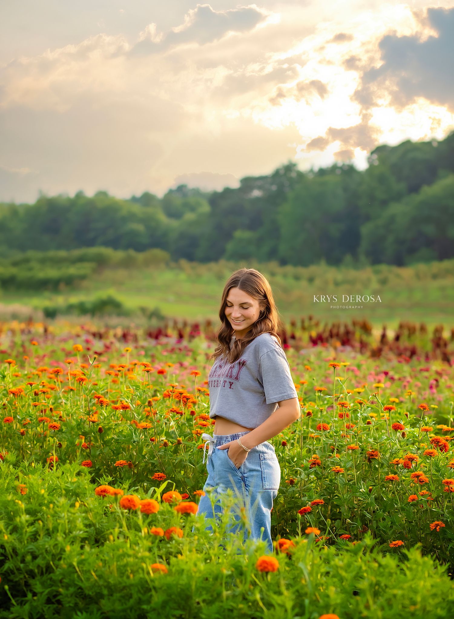 Young woman in gray tee and jeans smiles in a vibrant field of orange and red wildflowers at golden hour.