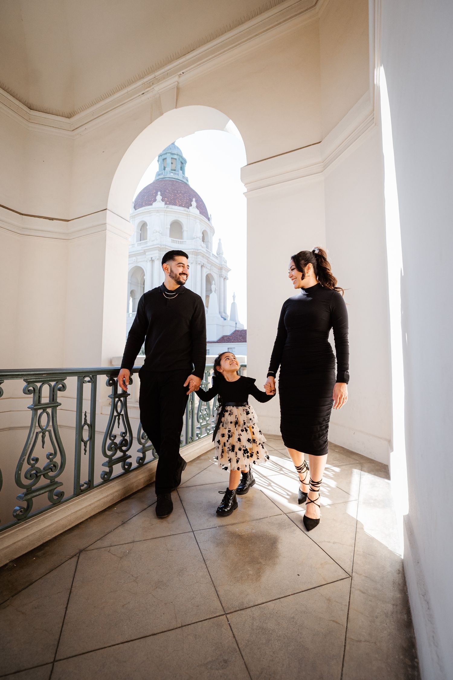 Family of three holding hands on a sunlit balcony with ornate railings and a domed Pasadena City Hall building backdrop.