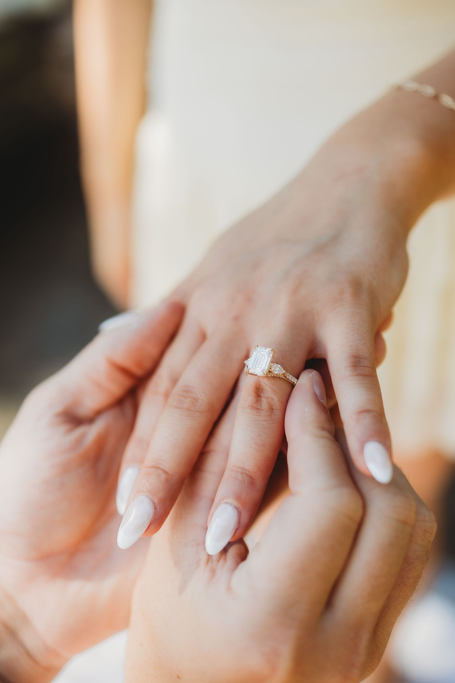 Close-up of hands placing a gold emerald-cut engagement ring on a bride's finger with white manicured nails.