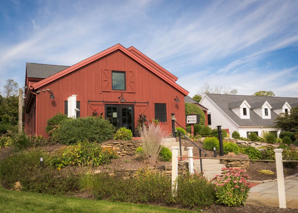 Red barn converted into a charming building surrounded by lush gardens and a white farmhouse under a blue sky.