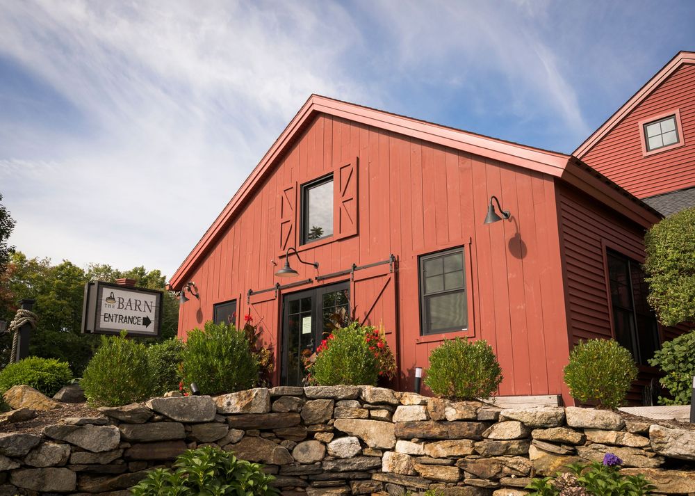 Red barn building with stone wall entrance, green shrubs, and a sign reading Barn Entrance under a partly cloudy sky.