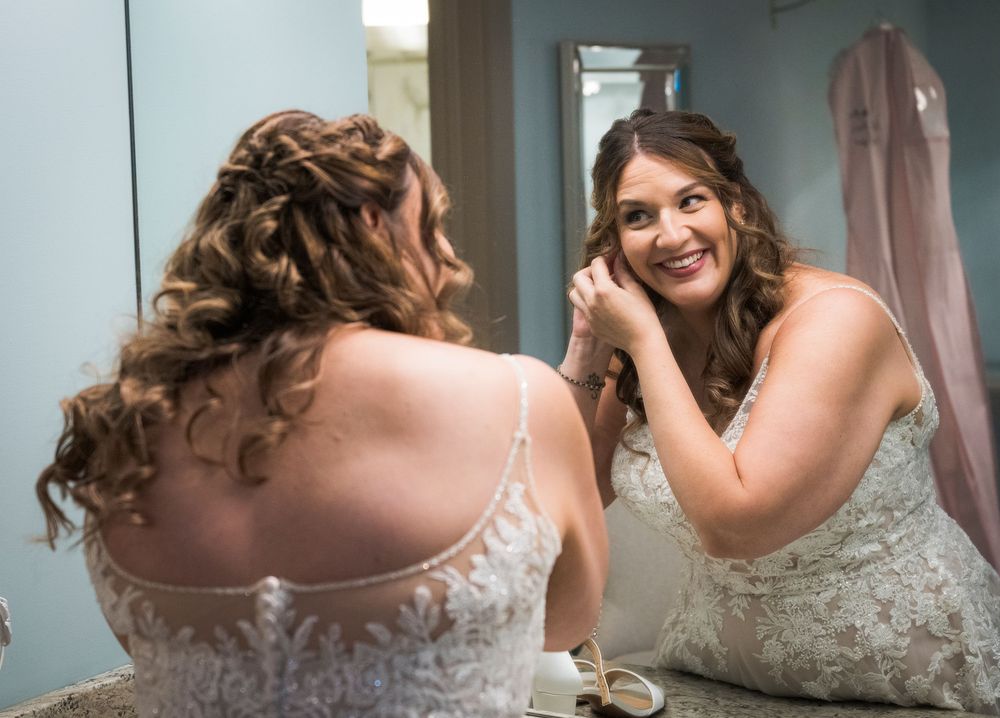 Bride in lace wedding dress smiling at her reflection in mirror while putting on earrings before ceremony.