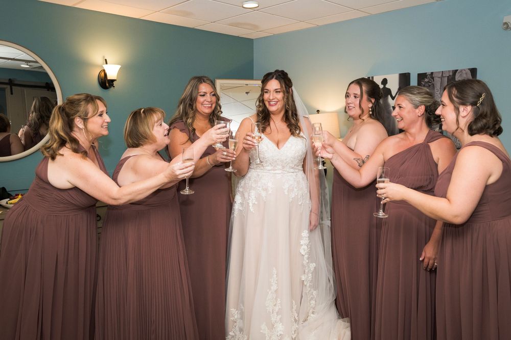Bride in white lace gown toasts champagne with six bridesmaids in mauve dresses during wedding preparations.