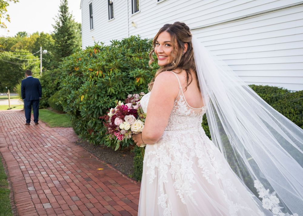 Bride in lace wedding dress and veil holds bouquet, smiling back at groom during first look on brick path.