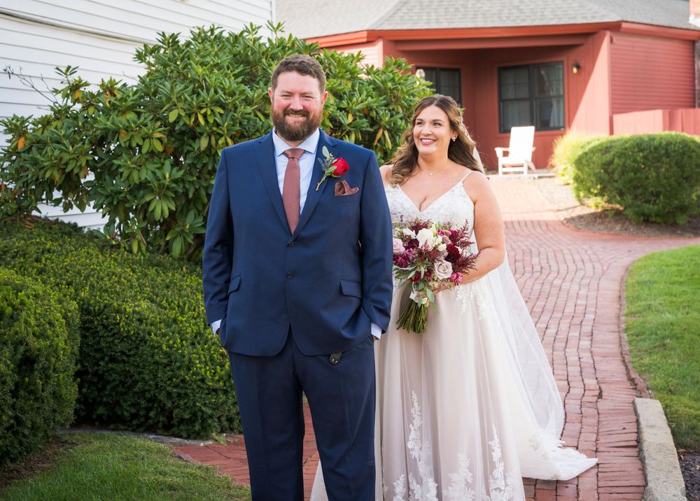 Bride in white gown holding bouquet stands with groom in navy suit outside red building on brick path.