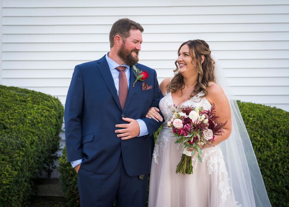 Bride and groom smiling at each other outdoors, she holds a burgundy bouquet, he wears a navy suit with floral boutonniere.