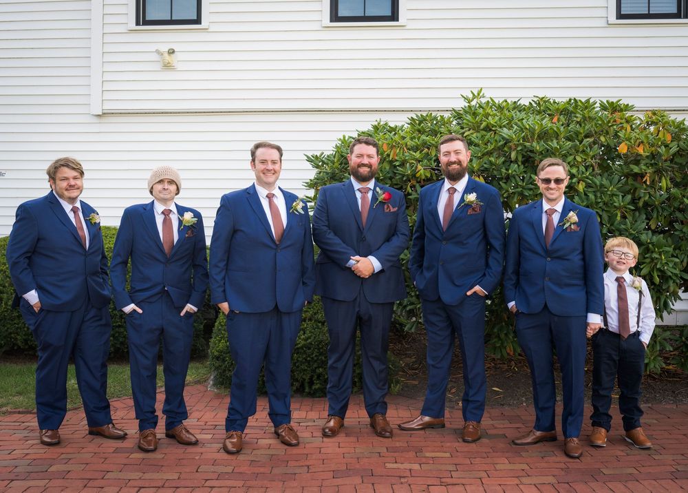 Seven groomsmen in navy blue suits with brown shoes pose together outdoors at a wedding ceremony.