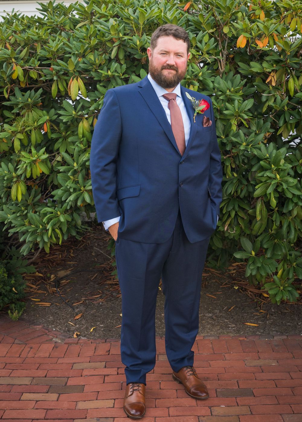 Man in navy blue suit with pink tie and boutonniere stands on brick path before lush green shrubs.