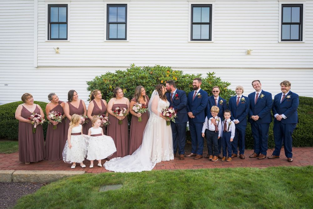 Wedding party poses outside white building, bridesmaids in mauve gowns, groomsmen in navy suits, with bride and groom centered.