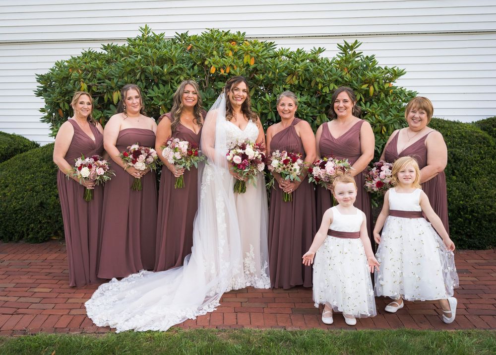 Bride with bridesmaids in mauve gowns and two flower girls in white dresses posing outdoors with floral bouquets.