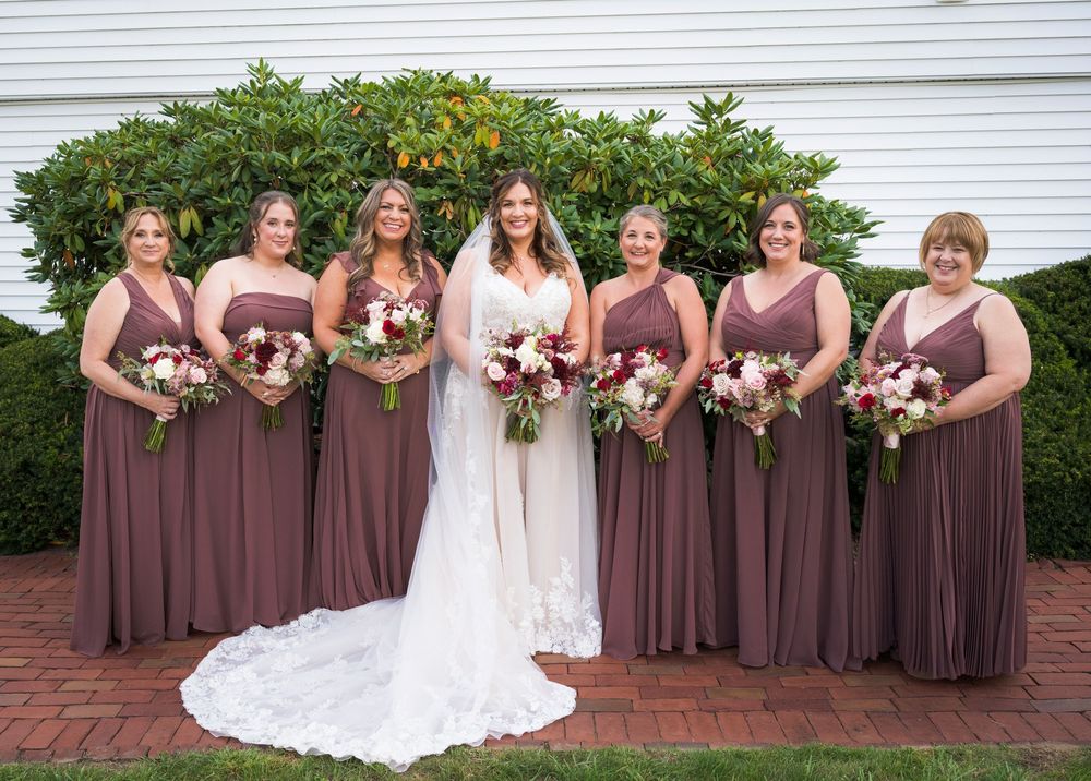 Bride and six bridesmaids in mauve gowns holding red and white bouquets posing outdoors before a wedding ceremony.