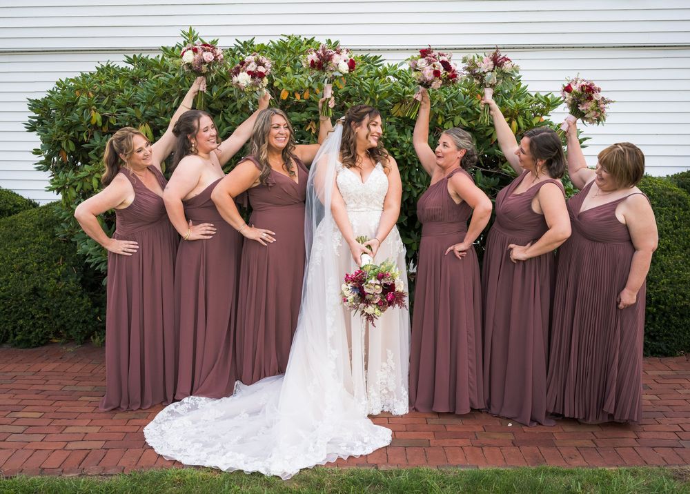 Bride with six bridesmaids in mauve dresses raising bouquets outside a white building.