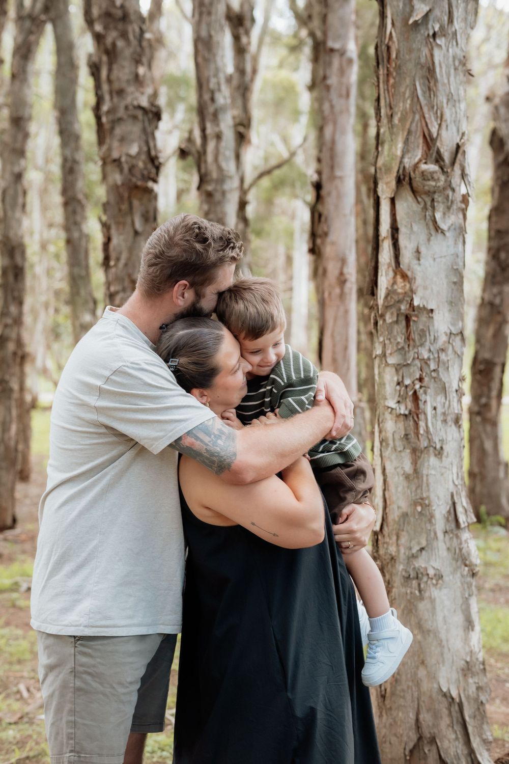 A family of three sharing a warm embrace among tall trees in a lush forest setting in Beaumont Hills