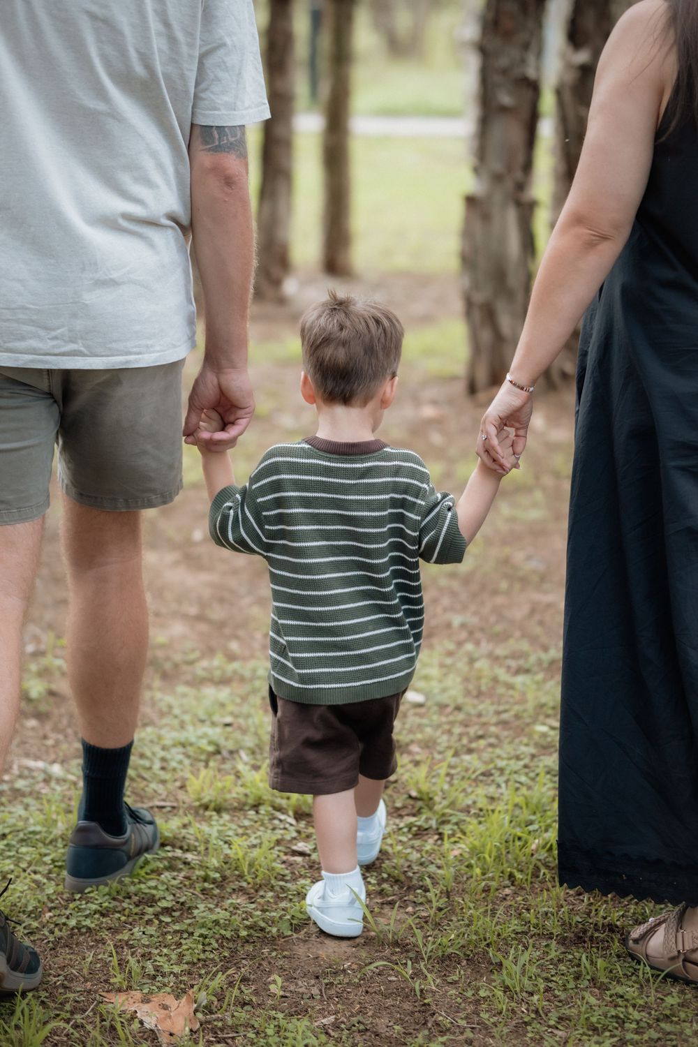 Young child holding hands with parents walking through a park, seen from behind, wearing striped sweater.