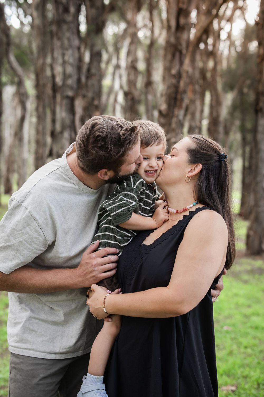 Parents kissing their young son on the cheeks in a lush green park, creating a joyful family moment outdoors.
