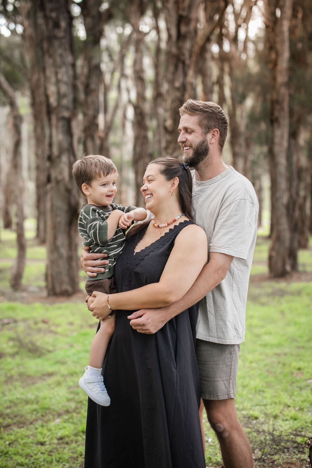 Happy family of three posing outdoors in a forest, pregnant mother in black dress held by smiling partner holding toddler son.