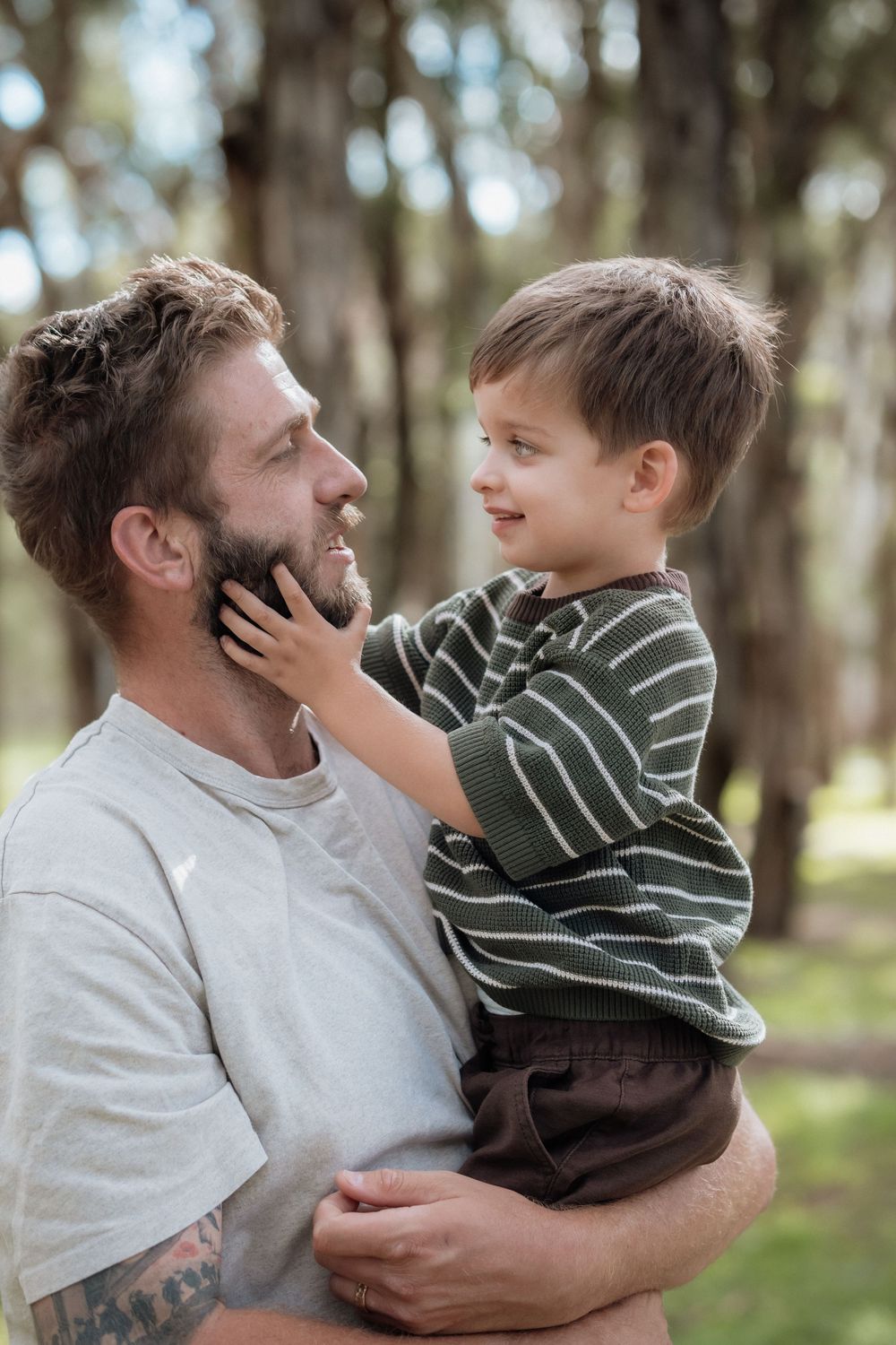 A father holds his young son in a park, as the boy lovingly touches his dad's bearded face.