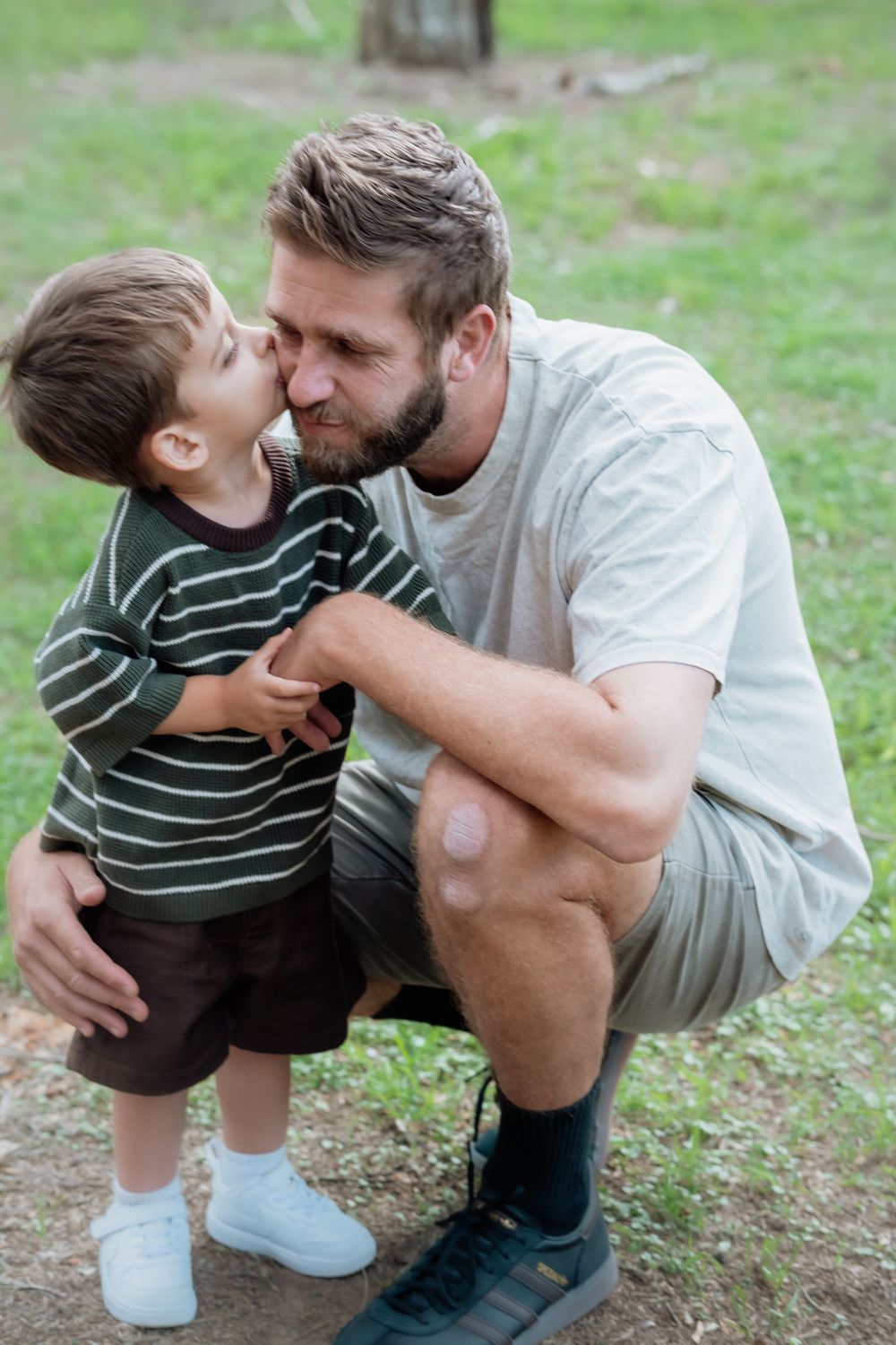 A father crouches down as his young son gives him a kiss on the cheek outdoors in a park.