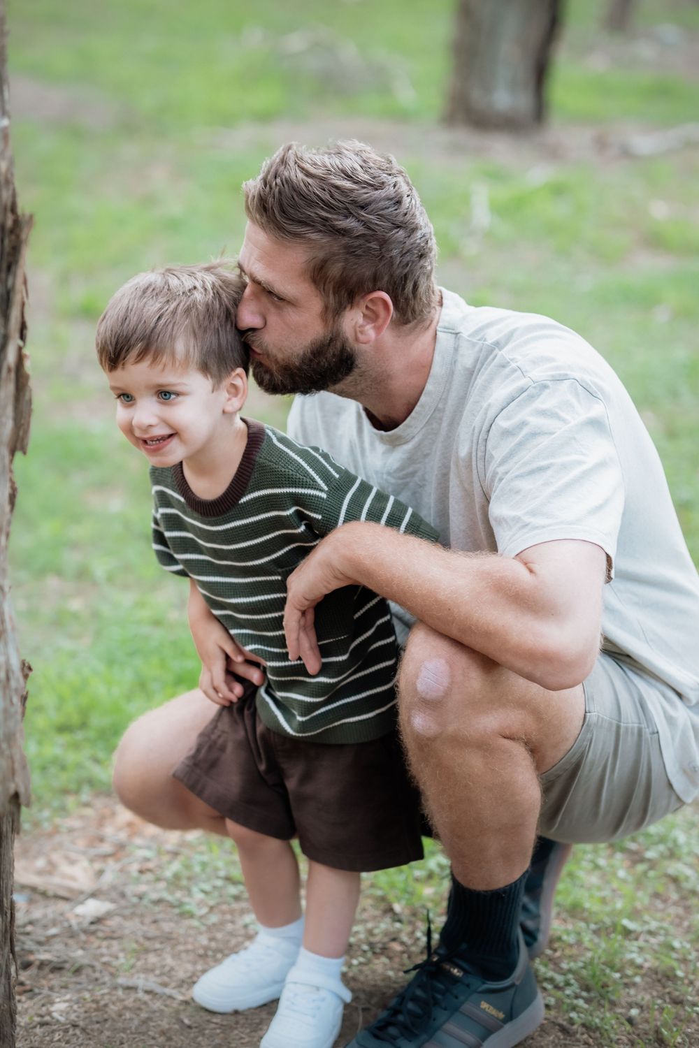 A bearded father crouches beside his young smiling son in a green park in the Hills District, Sydney, kissing his head affectionately.