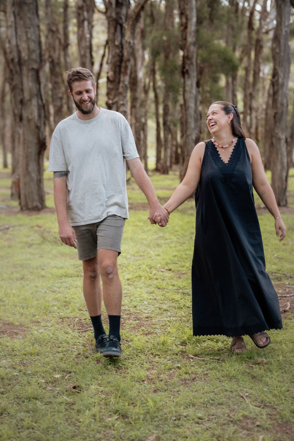 A happy couple walks hand-in-hand through a forest, man in gray tee and shorts, woman in black dress, smiling.