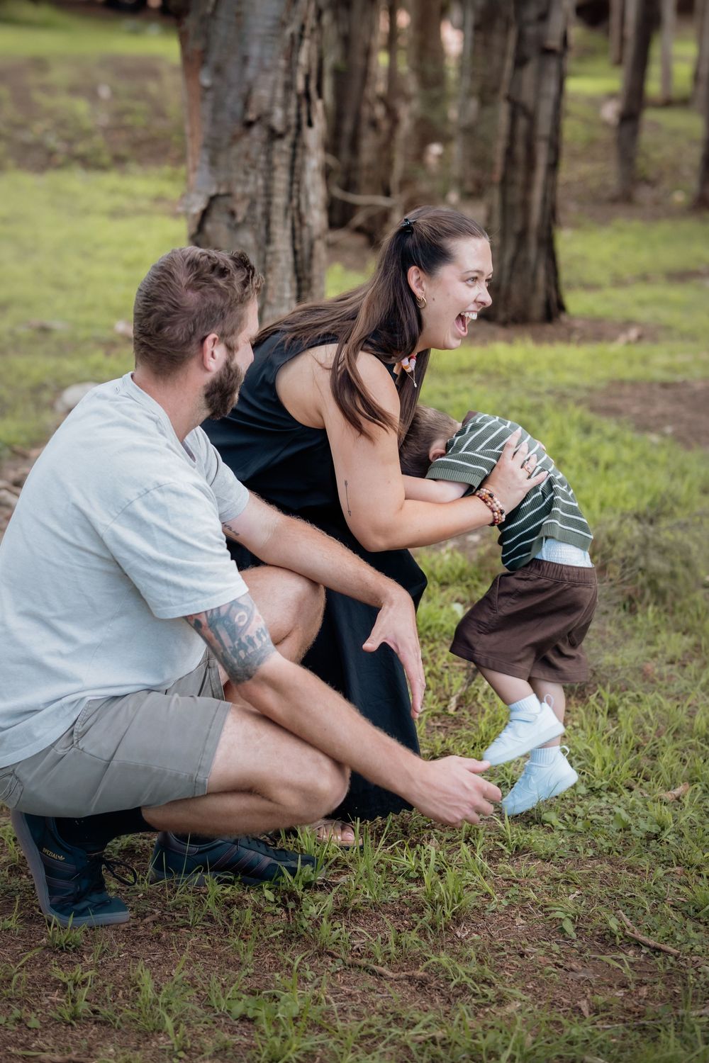 Young family laughing and playing together in a forest park, mother hugging toddler while father crouches beside them.