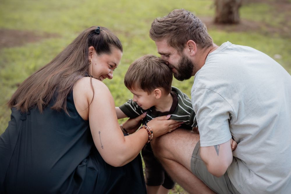 A happy family of three shares a tender moment outdoors, with parents leaning in to kiss their young son.