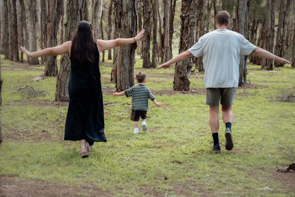 Family of three walking through a forest, parents and child with arms outstretched on green grass in Beaumont Hills