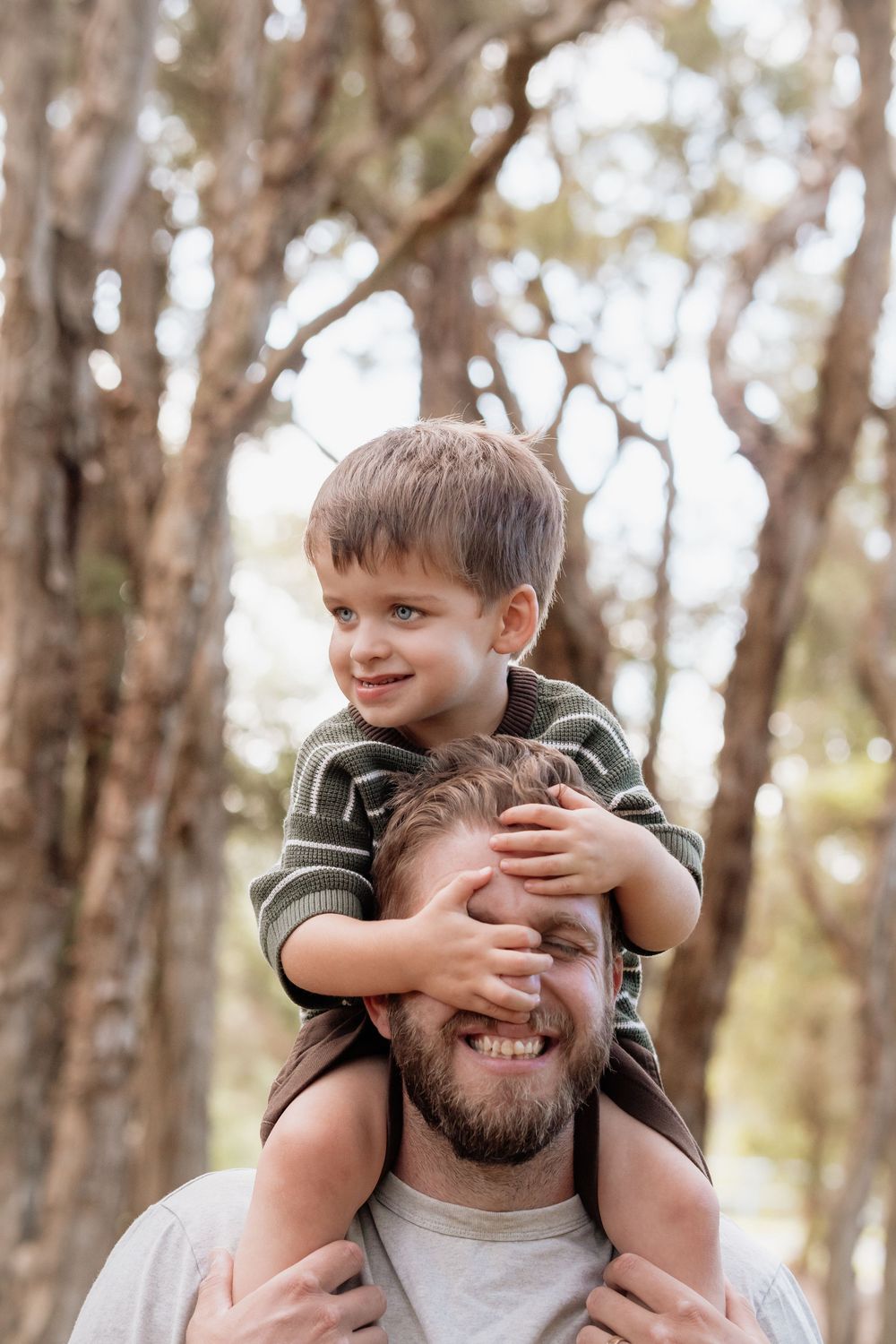 Happy father smiling as young boy sits on his shoulders covering his eyes in a sunlit forest setting.