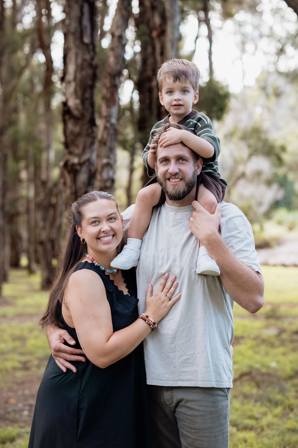 A happy family of three poses outdoors among tall trees, with a young boy sitting on his father's shoulders.
