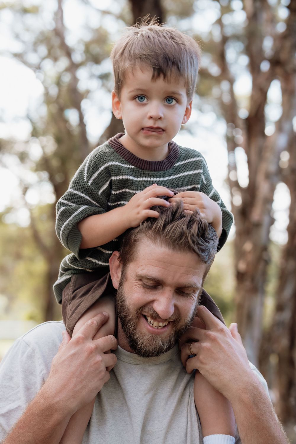 A bearded father smiles while carrying his young blue-eyed son on his shoulders outdoors among trees.
