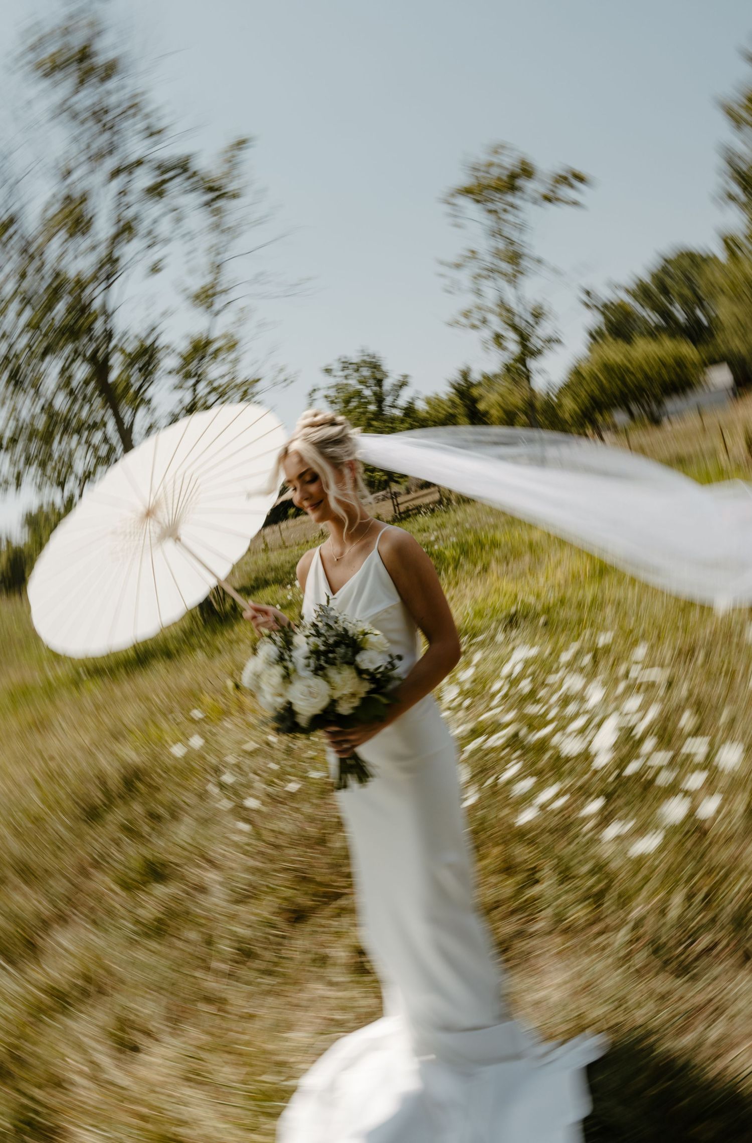 Bride in white gown holding bouquet and parasol, veil flowing in wind, standing in sunny green field.