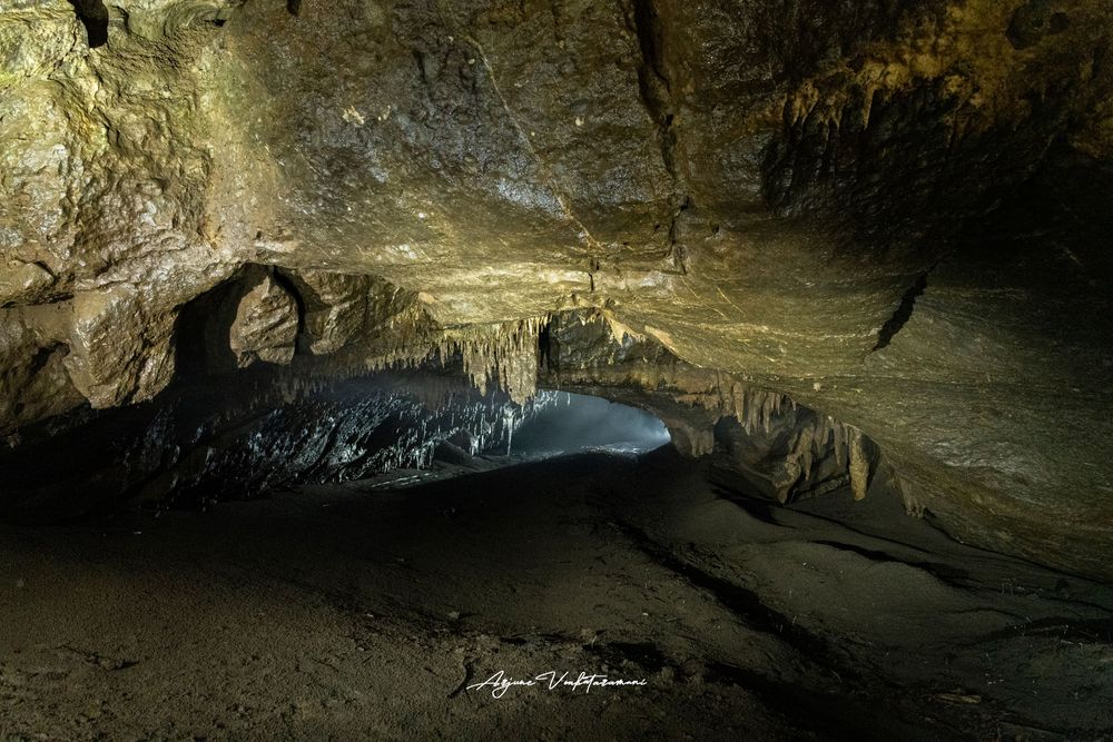 Dark cave interior with stalactites, rock formations, and a reflective pool illuminated by artificial light.