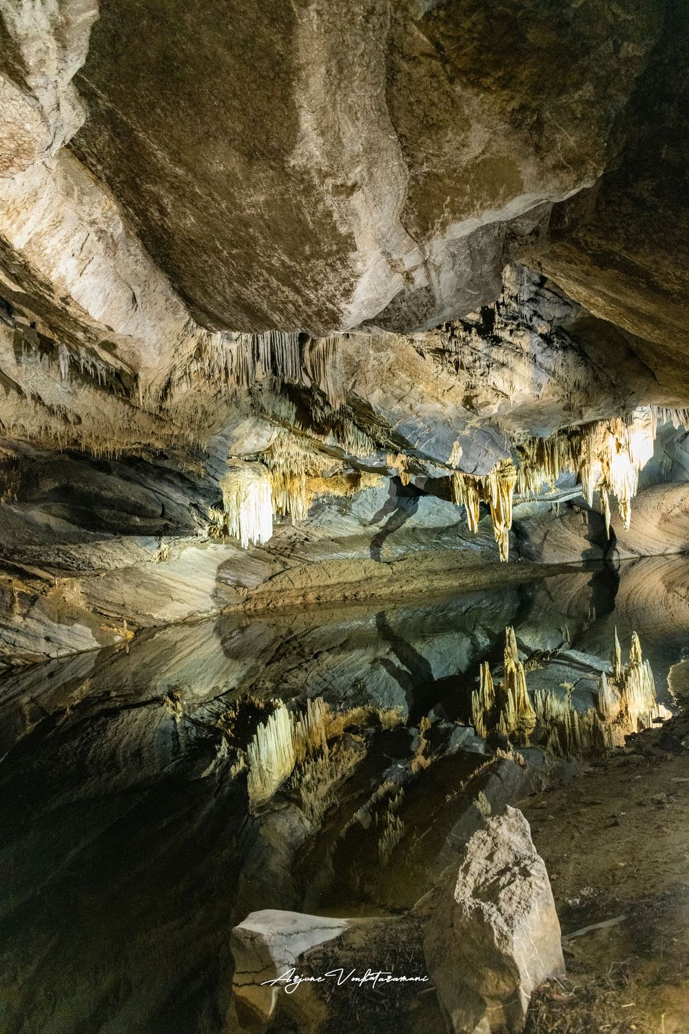Stunning cave interior with stalactites reflected in a still underground pool, illuminated by artificial lighting.