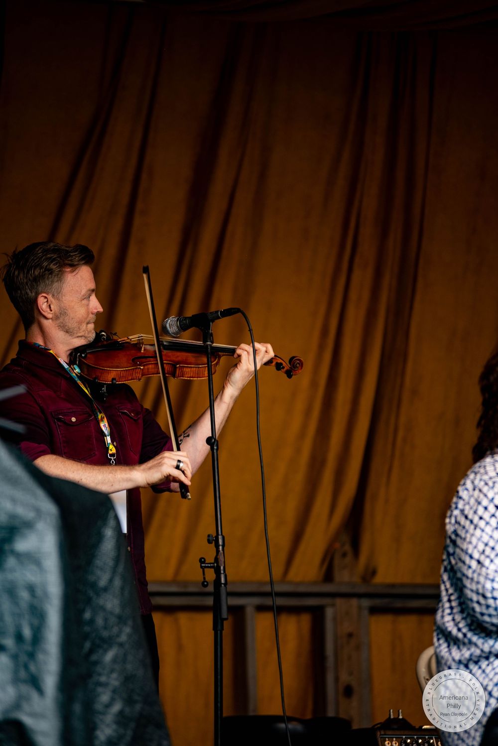 Man in red shirt playing violin on stage with microphone stand before a golden brown curtain backdrop.