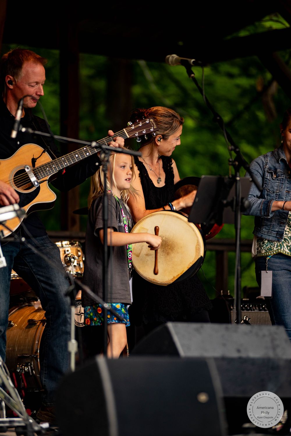 Young girl plays frame drum on stage with adult musicians including guitarist at outdoor music festival.